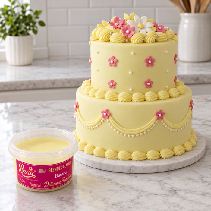 A yellow frosted two-tier cake decorated with piped swirls, pink and white flowers, and pearl-like dots, displayed on a marble board beside a tub of Beau Banana Blended Flavour frosting in a bright kitchen setting.