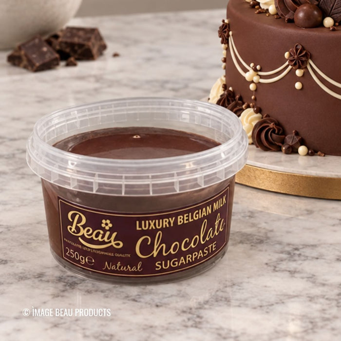 A tub of Beau's Luxury Belgian Milk Chocolate Sugarpaste on a marble surface, with a decorated chocolate cake and chocolate pieces blurred in the background.