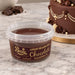 A tub of Beau's Luxury Belgian Milk Chocolate Sugarpaste on a marble surface, with a decorated chocolate cake and chocolate pieces blurred in the background.