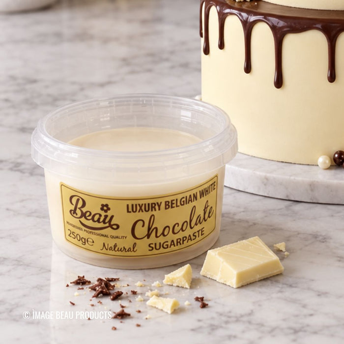 A tub of Beau Chocolate Sugarpaste labeled 'Luxury Belgian White' sits on a marble surface with chocolate pieces and crumbs around it, with a decorated cream cake blurred in the background.