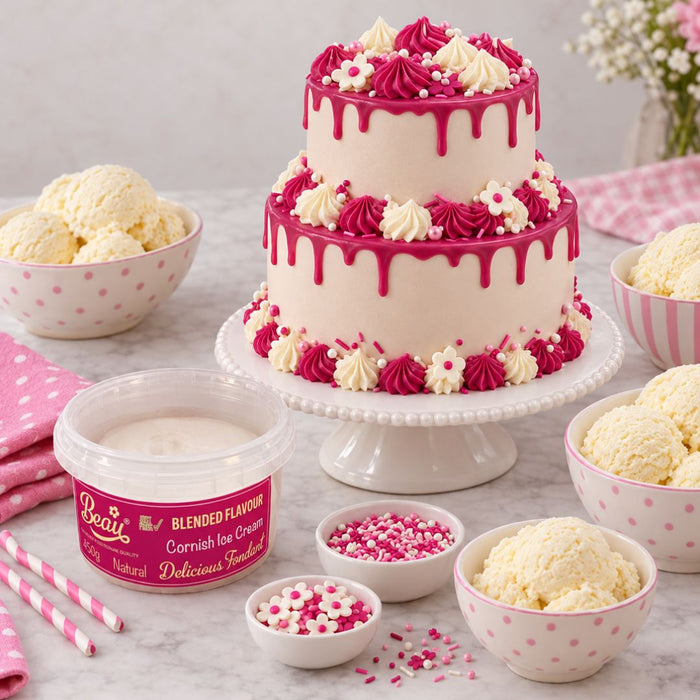 A two-tier decorated cake on a white pedestal stand, surrounded by bowls of vanilla ice cream, pink sprinkles, and a tub of Beau's Blended Flavour Cornish Ice Cream. The cake is decorated with white frosting, pink frosting, and white flowers.