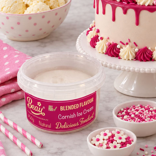 A tub of Beau's Blended Flavour Cornish Ice Cream placed on a marble surface, surrounded by pink and white dessert props including a decorated cake, bowls of sprinkles, ice cream, and striped straws.