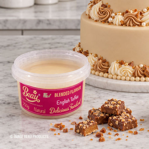 A tub of Beau's English Toffee in blended flavour, shown on a marble surface with a decorated cake and several pieces of toffee in front. The tub has a pink label, and the background includes a beige cake with cream and toffee decorations.