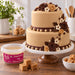 A two-tier beige cake decorated with dark and light brown chocolate roses, cream flowers, and beaded bands, displayed on a white cake stand. A tub of Beau's Blended Flavour Fudge is shown in the foreground on a wooden table, with loose and cubed fudge pieces nearby.