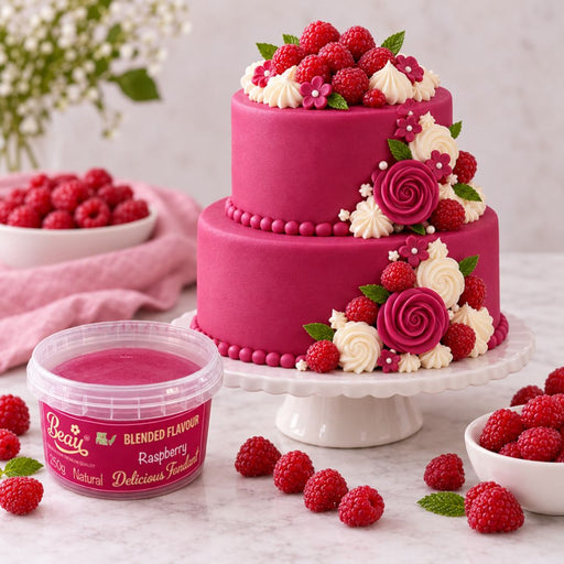 A two-tier pink cake decorated with fresh raspberries, white cream swirls, and pink rose decorations on a white cake stand, with a tub of Beau's Raspberry Blended Flavour frosting in the foreground and bowls of raspberries on a marble surface.