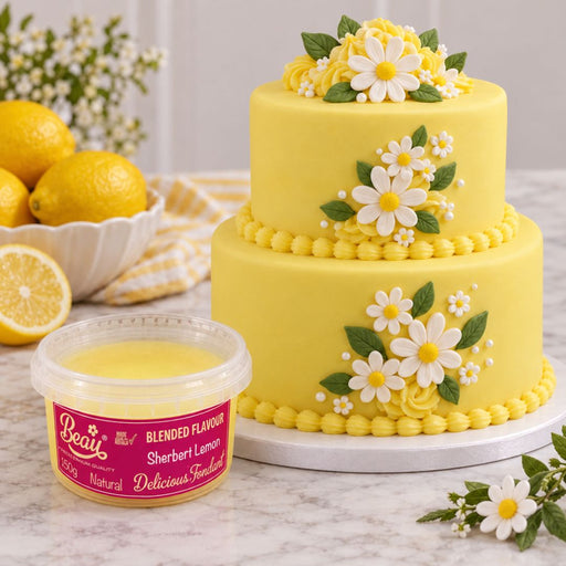 A yellow two-tier cake decorated with white daisy flowers and green leaves, shown on a marble surface beside a tub of Beau's Blended Flavour Sherbert Lemon frosting. Lemons and flowers are arranged in the background.