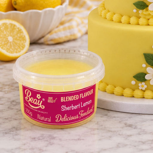 A tub of Beau's Sherbert Lemon Fondant in the Blended Flavour, shown in front of a decorated yellow cake and lemons on a marble surface.