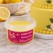 A tub of Beau's Sherbert Lemon Fondant in the Blended Flavour, shown in front of a decorated yellow cake and lemons on a marble surface.