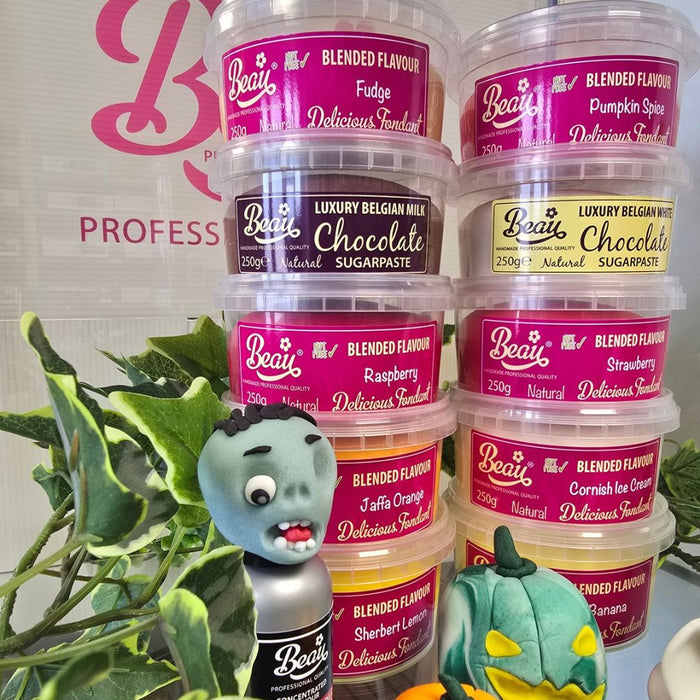 A stack of ten Beau's Professional blended flavor sugarpaste tubs in various colors, arranged with decorative props including a green plant, a small gray monster figurine, and a green pumpkin decoration against a white background with a pink logo.