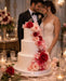 Wedding cake decorated with white flowerpaste with red and pink flowers in front of a couple in formal wedding attire.